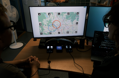 Two people wearing eye-tracking glasses look at a web map on a monitor framed by AprilTag markers, with coloured circles marking their gaze
